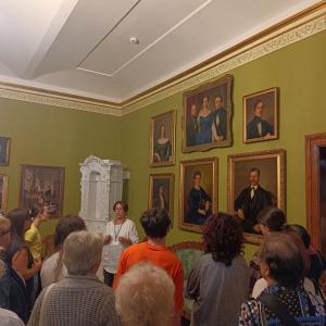 na zdjęciu grupa ludzi zwrócona w stronę P. Teresy D. Bujarek, w tle obrazy P.M.Bohuna wiszące na zielonej ścianie, w tle duży, biały piec