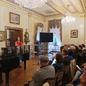 Na zdjęciu widoczna jest kobieta w eleganckiej czerwonej sukience, uśmiechająca się i witająca gości, trzymając w ręku mikrofon. W tle znajduje się czarny fortepian, a goście siedzą na krzesłach w stylowym salonie muzycznym, który emanuje atmosferą klasyc