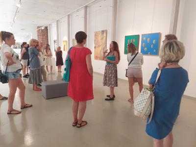 ludzie zgromadzeni wokół P. Agaty Smalcerz, słuchający jej oprowadzania, w tle na ścianie - wiszące obrazy przedstawiające 5. Bielski Festiwal Sztuk Wizualnych