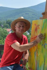 Zdjęcie przedstawiające artystę J.Motylskiego. Mężczyzna jest w średnim wieku, ubrany w czerwoną podkoszulkę, ciemne, niebieskie jeansy i słomiany kapelusz. Artysta w prawej dłoni trzyma czerwony pędzel, który znajduje się na obrazie. Obraz jest w kolorac