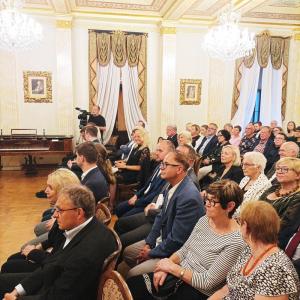 Salon muzyczny jest wypełniony po brzegi zgromadzoną publicznością, która z uwagą i uśmiechem śledzi występ muzyków. Wszyscy z wyraźnym skupieniem wpatrują się w artystów, którzy oddają się grze. W tle, w pobliżu, widać kamerzystę, który prowadzi transmis