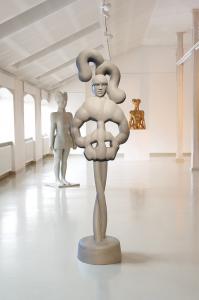 Widok ogólny rzeźb Agaty Agatowskiej w białej przestrzeni galerii w Starej Fabryce (poddasze). Na pierwszym planie znajduje się fantazyjna rzeźba, całopostaciowa – w ogólnym wrażeniu przypominającym postać baletnicy w układzie ze skrzyżowanymi nogami i rę