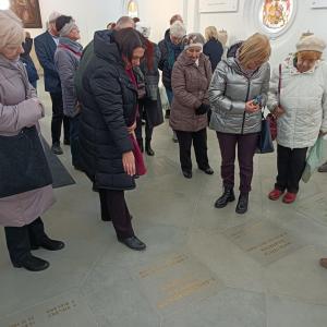 grupa ludzi stojąca za P. Bożeną Chorąży, która prawą ręką pokazuje miejsce krypt rodziny Sułkowskich, wszyscy mają głowy skierowane w stronę szarej posadzki podłogowej