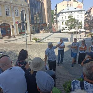 Na środku zdjęcia znajduje się P. Bogusław Chorąży ubrany w białą koszulę z krótkim rękawem i czarne, długie spodnie. P. Bogusław w lewej dłoni, skierowanej do góry- trzyma grafikę. Przed nim zgromadzona jest spora grupa ludzi w różnym przedziale wiekowym